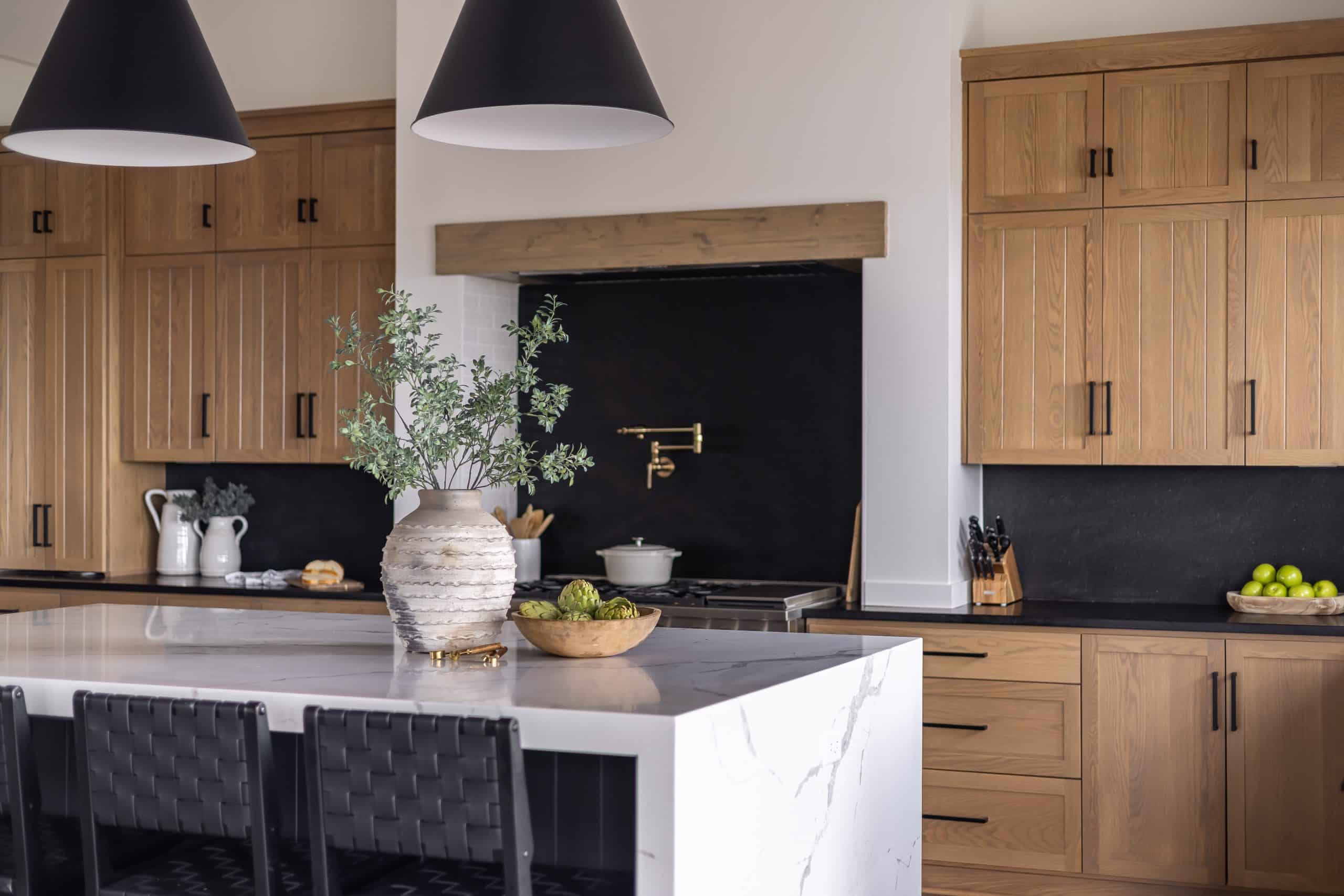 Classic cabinets, modern marble countertop that extends to floor. Classic vase and thoughtful greenery, with dark elements in light shades, backsplash, and chairs. Dark accents to compliment wood cabinets. Classic gold hardware. 3D Rendering